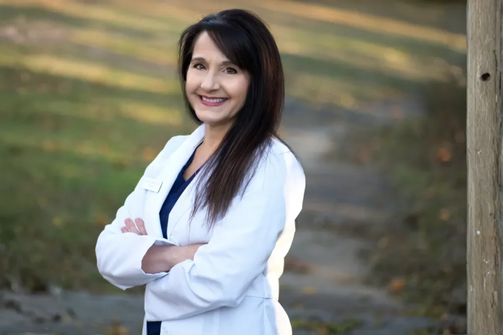 Beckman | Dry Eye Treatment Smiling woman in a white lab coat stands with arms crossed outdoors on a park path near a wooden post.