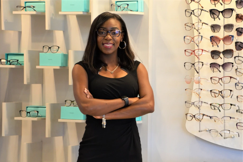 Woman in a black dress with arms crossed stands in an eyewear store, shelves of glasses and turquoise boxes behind her.