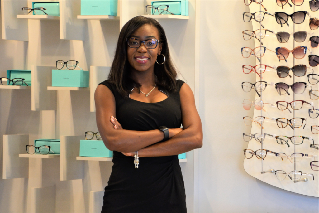 Smiling woman with glasses standing in a sunglasses store, arms crossed.